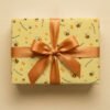Pale yellow wrapping paper with repeating pattern of cartoon bees, honeycomb shapes, and "Happy Birthday" text in navy blue, wrapped as a gift box with a golden-bronze satin ribbon bow on a neutral background.