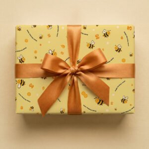 Pale yellow wrapping paper with repeating pattern of cartoon bees, honeycomb shapes, and "Happy Birthday" text in navy blue, wrapped as a gift box with a golden-bronze satin ribbon bow on a neutral background.