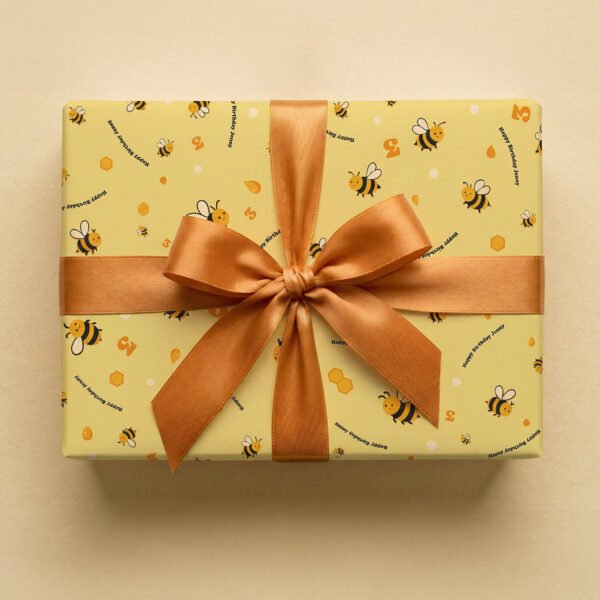 Pale yellow wrapping paper with repeating pattern of cartoon bees, honeycomb shapes, and "Happy Birthday" text in navy blue, wrapped as a gift box with a golden-bronze satin ribbon bow on a neutral background.