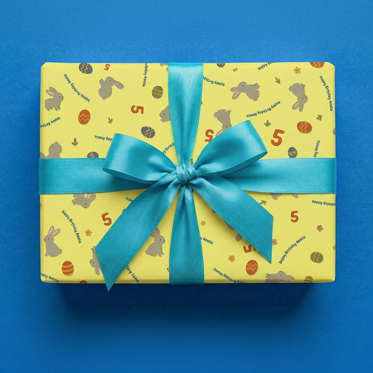 Bright yellow wrapping paper with Easter-themed pattern wrapped as a gift box with a turquoise satin ribbon bow, featuring cartoon rabbits in gray with pink inner ears, decorated Easter eggs in red, blue, and yellow with various patterns, age number "5", small flowers, and personalized text on a blue background.