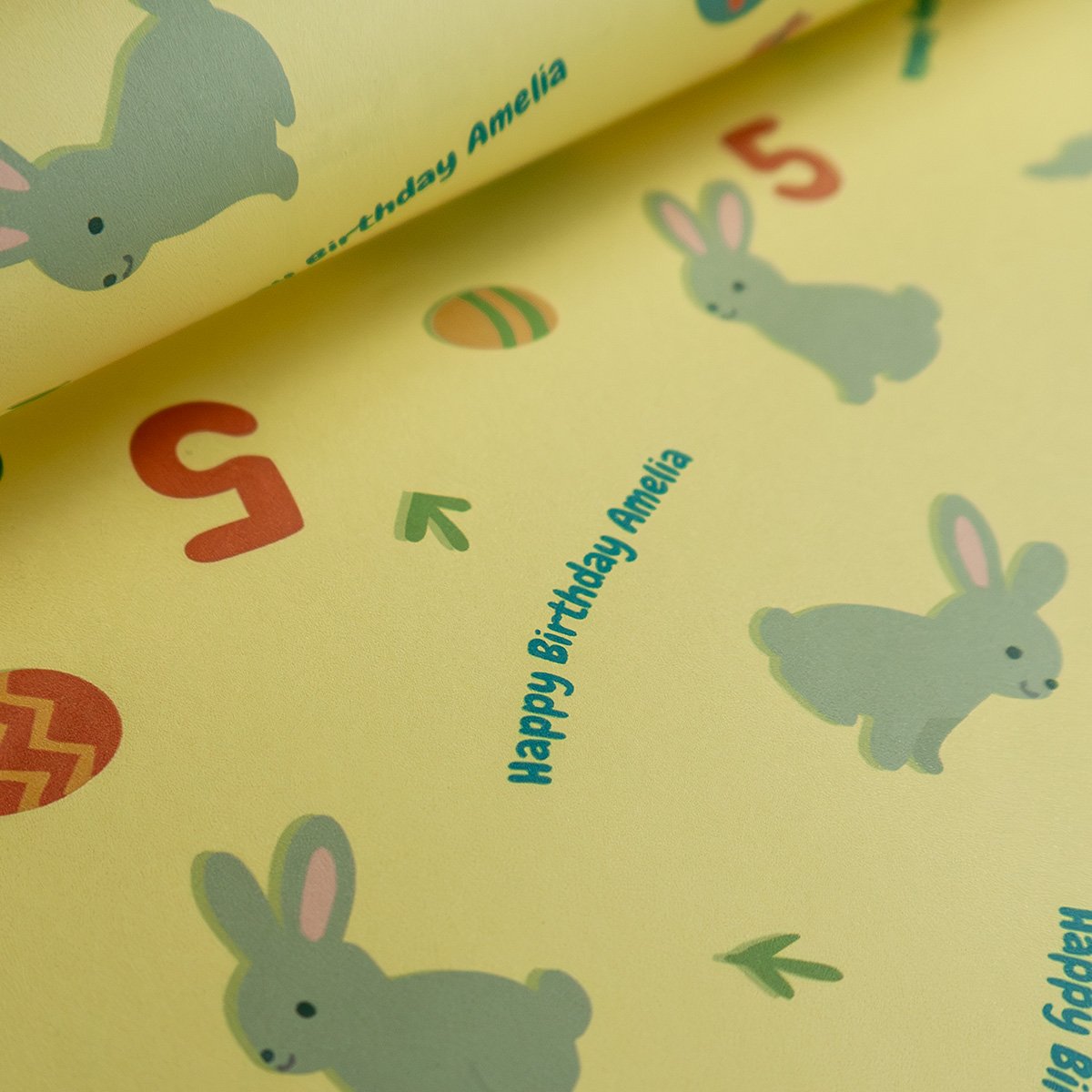 Close-up of rolled wrapping paper showing personalized text "Happy Birthday Amelia" in teal blue alongside gray cartoon bunny characters with pink inner ears, red Easter eggs with chevron patterns, and small flower and arrow decorative elements on the bright yellow background.