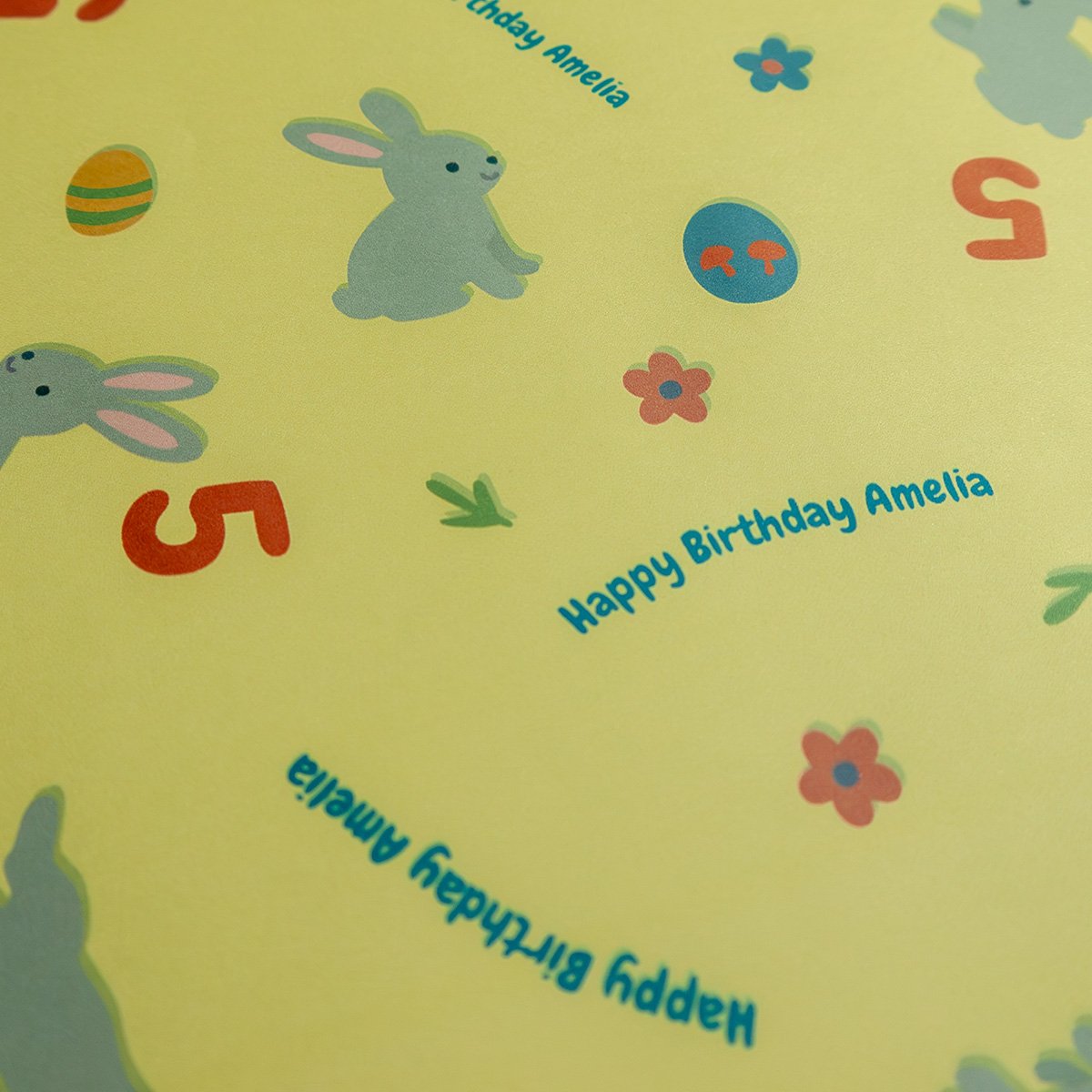 Detailed close-up of wrapping paper showing personalized text "Happy Birthday Amelia" in teal blue, gray cartoon rabbits with characteristic pink ears and simple dot eyes, decorated Easter eggs in various colors and patterns, red age numbers "5", and small coral and blue flowers on the yellow stock.