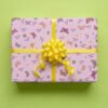 Pale pink wrapping paper with butterfly and flower pattern wrapped as a gift box with a bright yellow satin ribbon bow and streamers, featuring colorful illustrated butterflies in orange, pink, purple, and yellow, small daisy flowers in various colors, and personalized "Best Wishes Nicola" text on a lime green background.