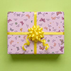Pale pink wrapping paper with butterfly and flower pattern wrapped as a gift box with a bright yellow satin ribbon bow and streamers, featuring colorful illustrated butterflies in orange, pink, purple, and yellow, small daisy flowers in various colors, and personalized "Best Wishes Nicola" text on a lime green background.